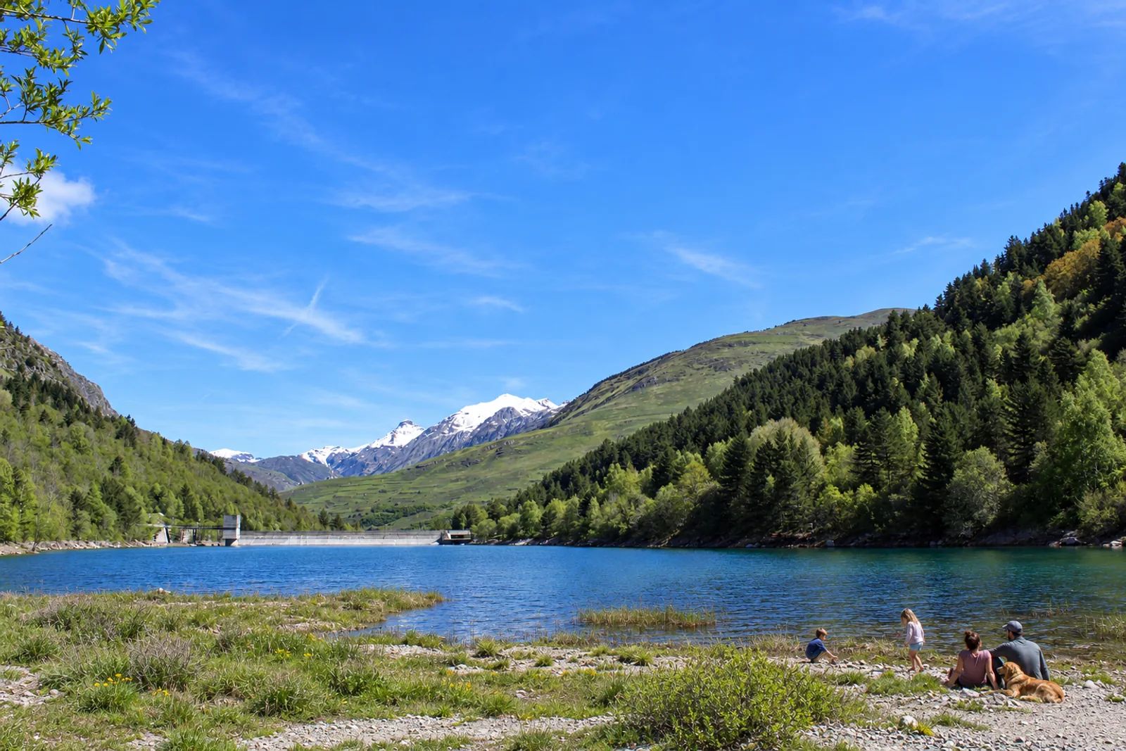 Familia con perro junto al agua de la presa de Aiguamòg, con la presa y el valle al fondo
